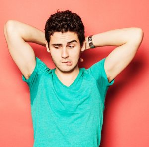 Young man biting lip while looking at sweaty armpit against red background