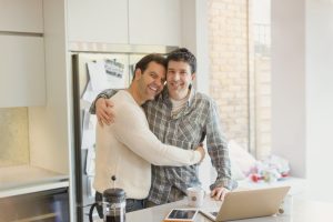 Portrait affectionate male gay couple hugging at laptop in kitchen