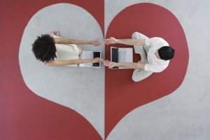 Man and woman sitting with laptops on large heart, arms out, touching hands, overhead view