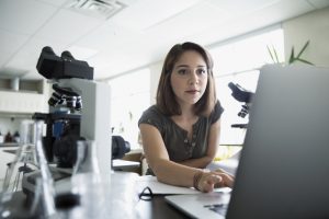 Focused college student using laptop near microscopes in science laboratory