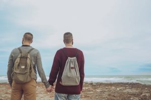 Back view of young gay couple with backpacks standing hand in hand on the beach looking at the sea