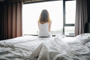 Portrait of depressed woman sitting alone on bed, looking to outside the window.