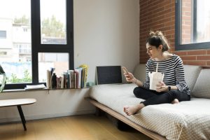 Young woman sitting on couch at home having Asian takeaway food and using cell phone
