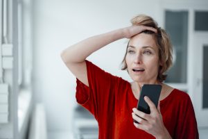 Businesswoman with mouth open and hand in hair holding smart phone in office