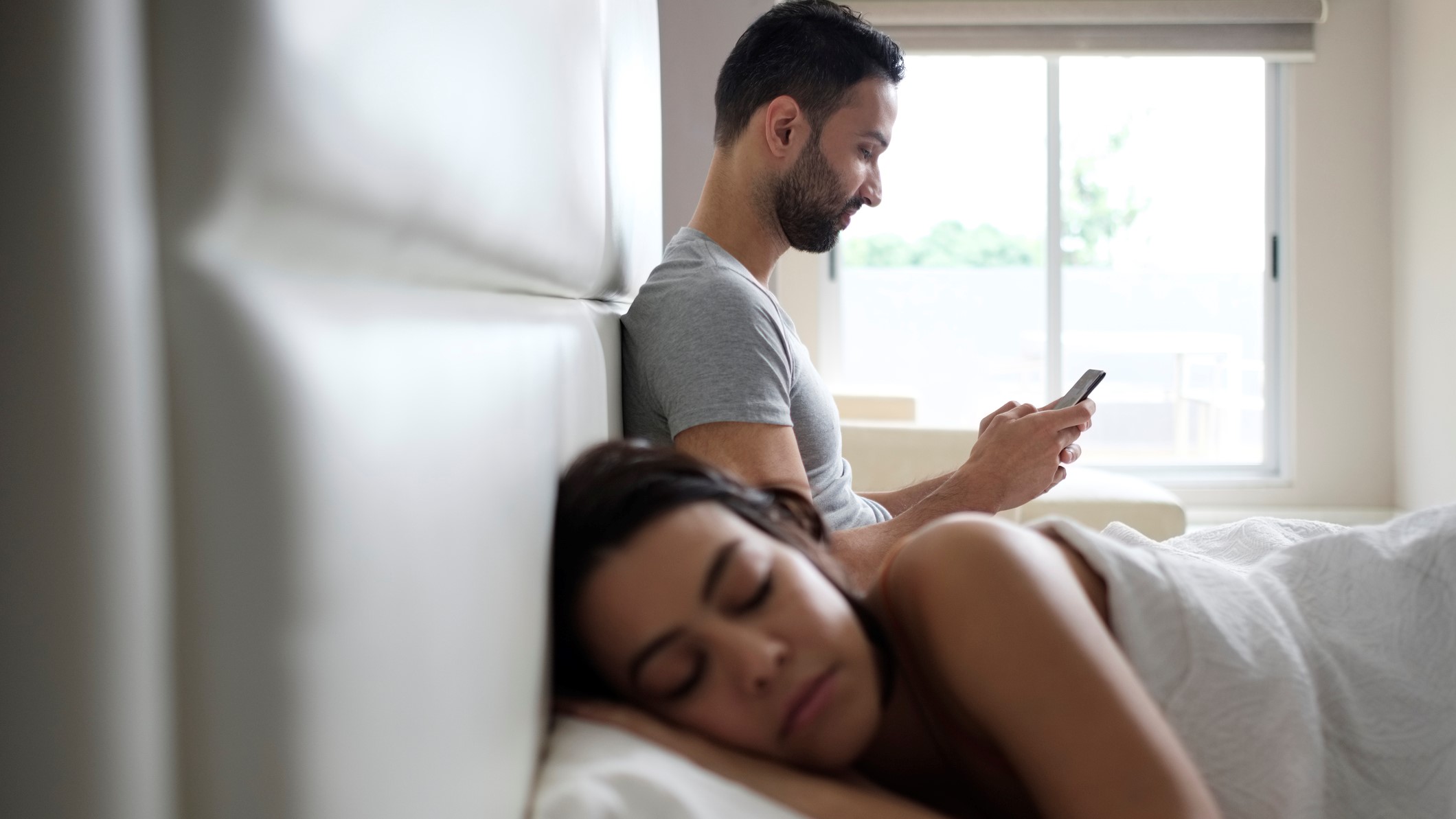 Un chico consulta algo en su móvil, mientras su chica duerme al otro lado de la cama.