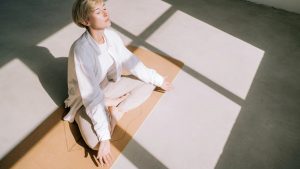Beautiful authentic woman with short blond hair is meditating sitting in lotus position on yoga mat in front of a window. She is wearing a light-colored casual clothing. Concept of relaxation exercises