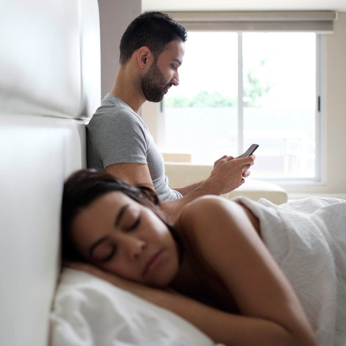 Un chico sentado en la cama mira el móvil y al lado está su chica durmiendo.