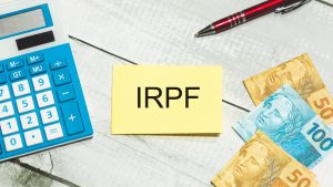 The initials IRPF do Imposto de Renda da Pessoa Física in Brazilian Portuguese written on a piece of paper on a light wooden table. A calculator, a pen and Brazilian Real banknotes in the composition.