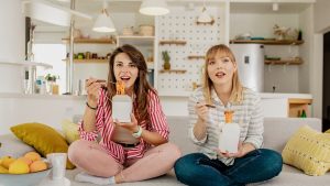 Two female friends are eating pasta at home and watching TV show