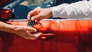 Dealer Giving Car Keys To The New Owner. Close-Up Of Hands On The Background Of Red Automobile