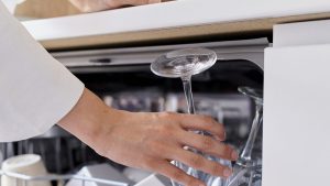 a white woman with long hair in purple pants and white blouse puts crockery to wash in the dishwasher