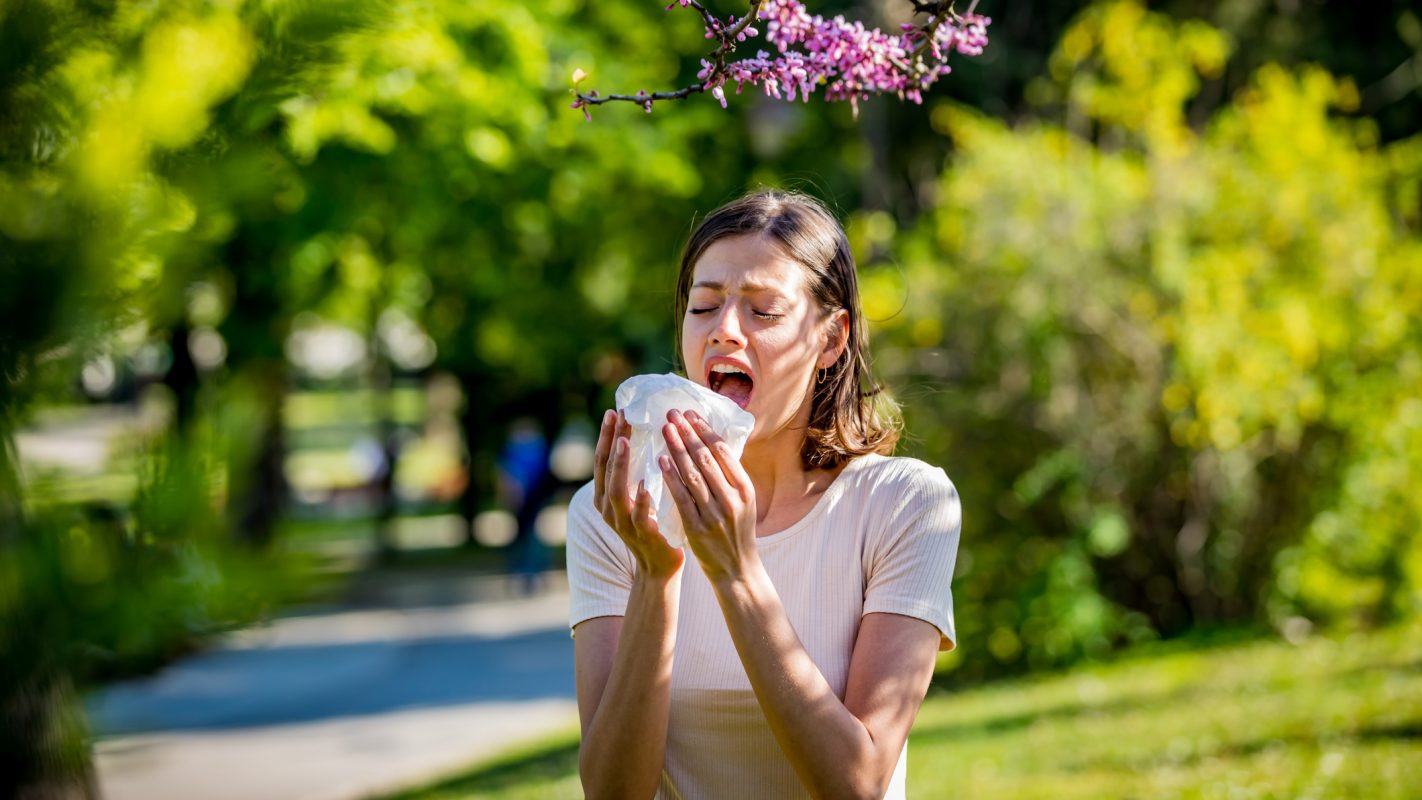 Una chica estornudando en plena calle al lado de la vegetación.