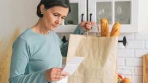 Senior adult female worried while looking at receipt from grocery store. Rise in price of products concept. Buying consumer goods during inflation. Facing economic downturn. Kitchen unit and paper bag on background, horizontal. closeup