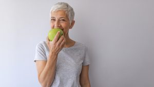 Beautiful senior woman over grey wall eating green apple.