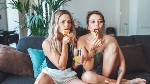 Two young concentrating and serious women watching fixedly TV, eating pizza and drinking lemonade indoors