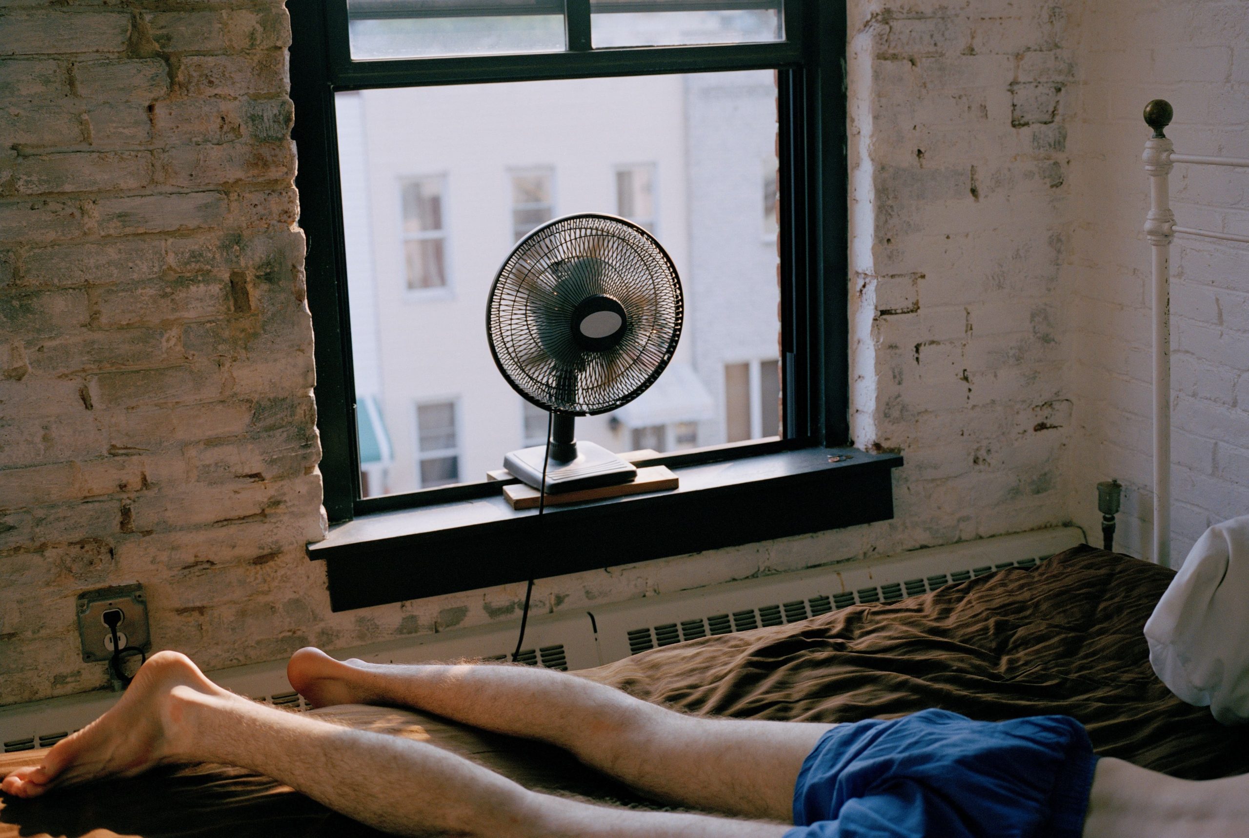 Un chico tumbado en la cama mientras tiene el ventilador encendido.