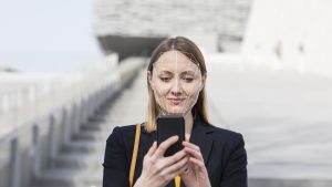 Businesswoman unlocking mobile phone with face recognition technology while standing outdoors