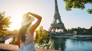Young woman tourist in sun hat and white dress standing in front of Eiffel Tower in Paris at sunset. Travel in France, tourism concept