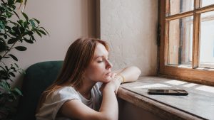 Upset redhead teen girl sitting by window looking at phone waiting call or message