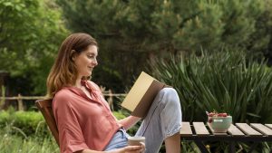 Woman with coffee mug reading book while sitting on chair in garden