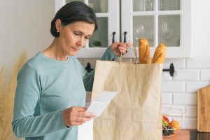 Senior adult female worried while looking at receipt from grocery store. Rise in price of products concept. Buying consumer goods during inflation. Facing economic downturn. Kitchen unit and paper bag on background, horizontal. closeup