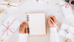 From above anonymous woman in knitted sweater making New Year's goals in blank planner near Christmas gifts and confetti on gray background. New Year's celebration concept