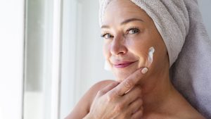 Smiling mature woman applying moisturizer on face at home