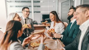 Small group of happy colleagues in formal wear chatting and eating pizza together for lunch. Alone we can do so little, together we can do so much.