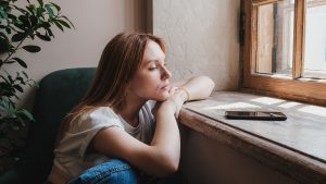 Upset redhead teen girl sitting by window looking at phone waiting call or message