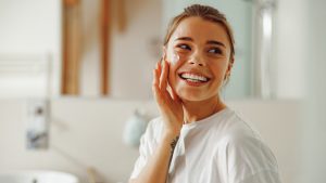 Young beautiful woman taking care of skin by applying moisturizer cream in bathroom